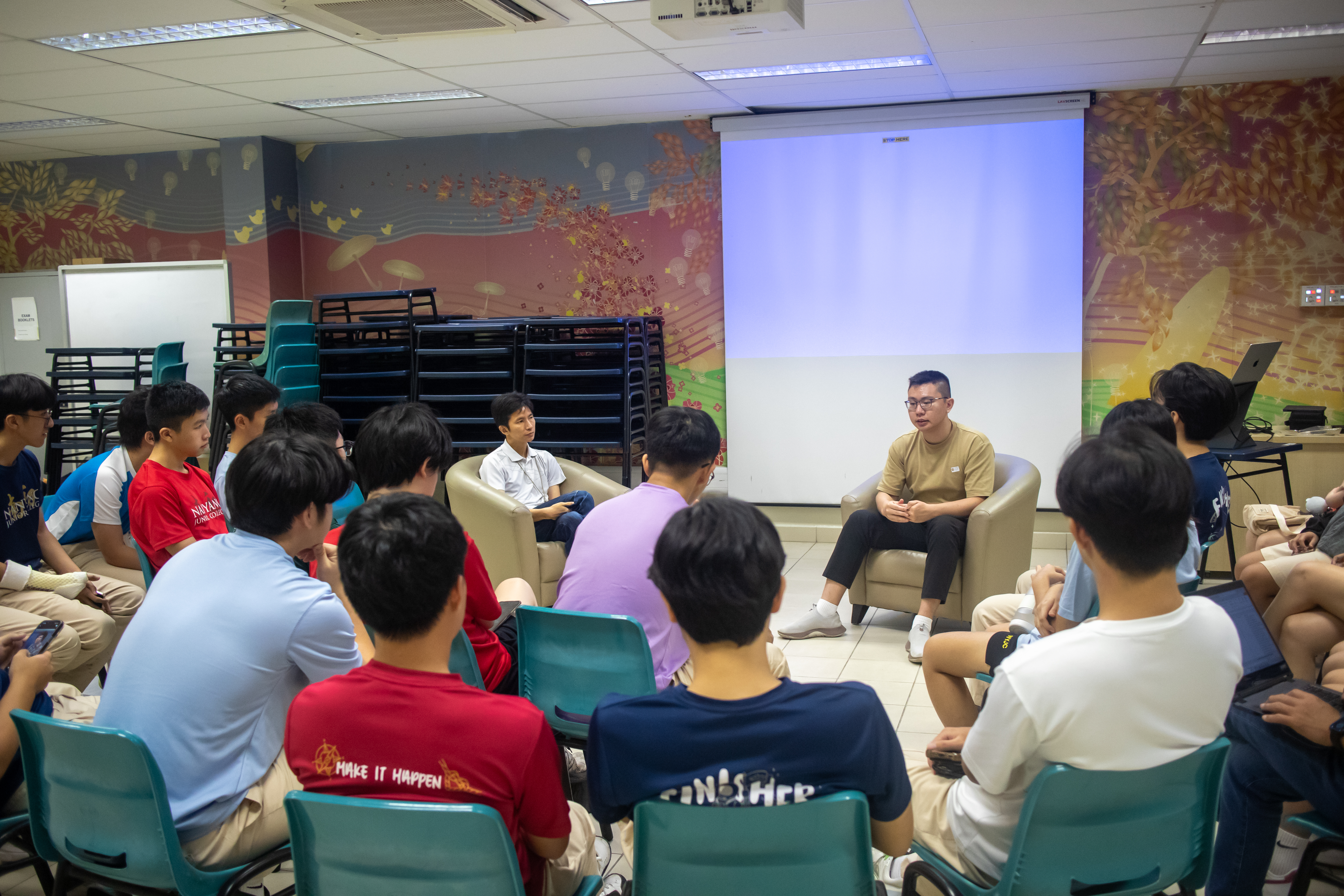 Huikang seated in a casual fireside chat setup, engaging with a group of students arranged in a semi-circle within a vibrant room featuring mural-style wall art.